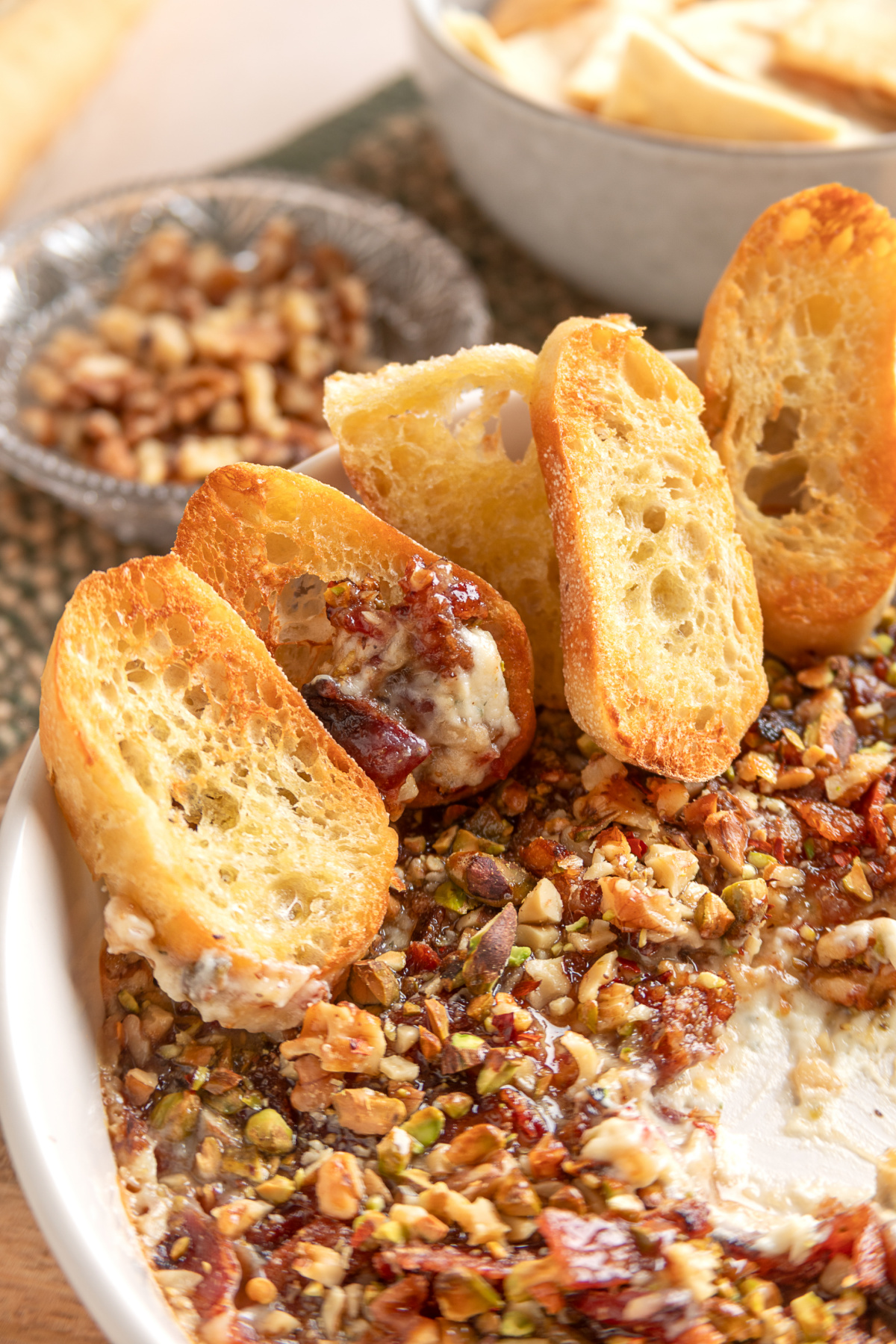 Toasted baguette slices standing in a warm baked Boursin dip topped with bacon, fig preserves, pistachios, and walnuts.