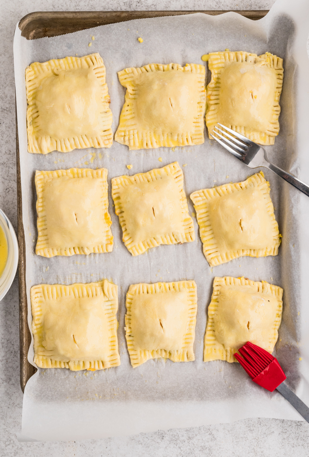 Unbaked puff pastry breakfast pastries sealed with fork-crimped edges and brushed with egg wash on a parchment-lined baking sheet.