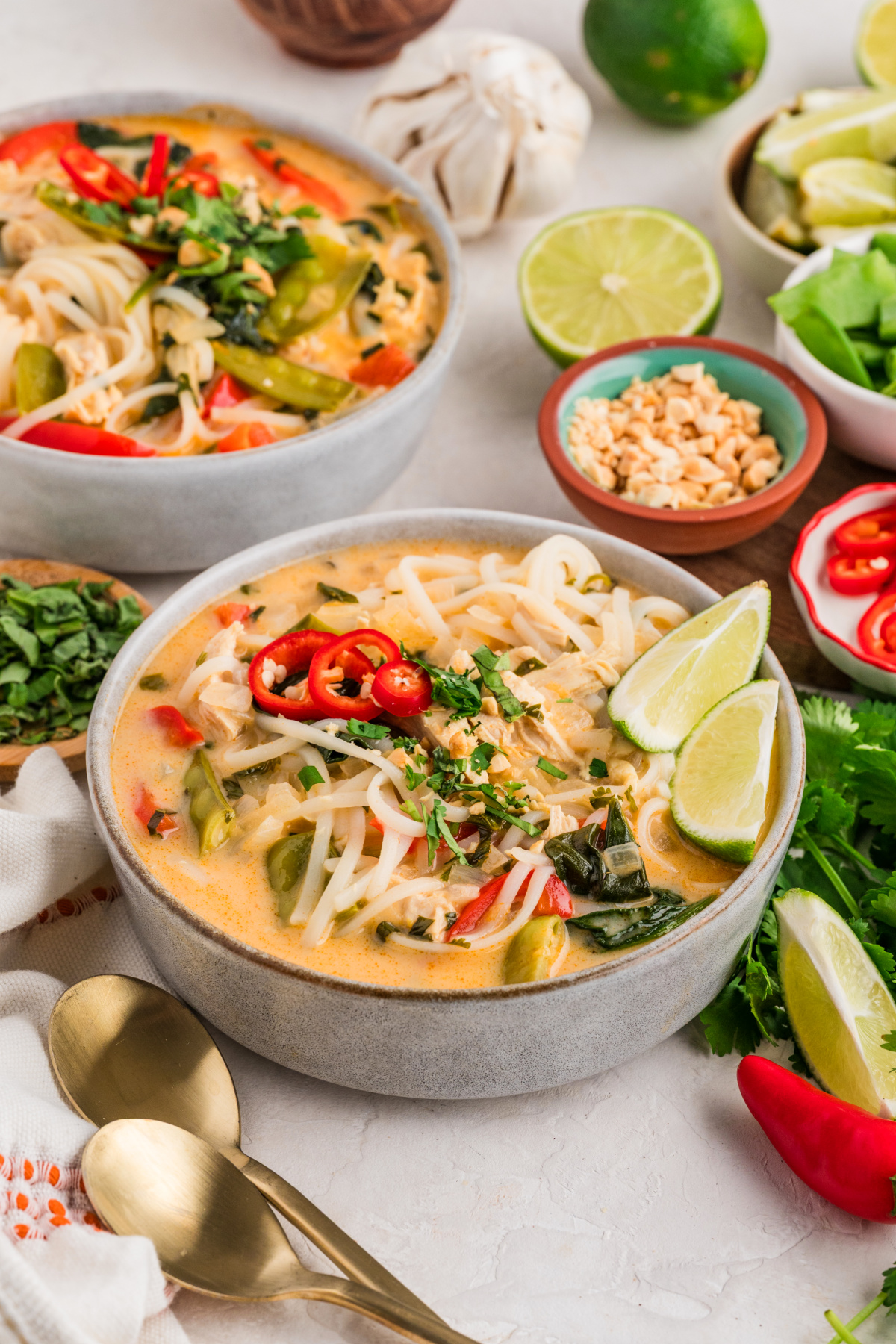 Chicken curry soup with coconut milk served in bowls, filled with rice noodles, shredded chicken, snow peas, red bell pepper, fresh herbs, sliced chili, and lime wedges.