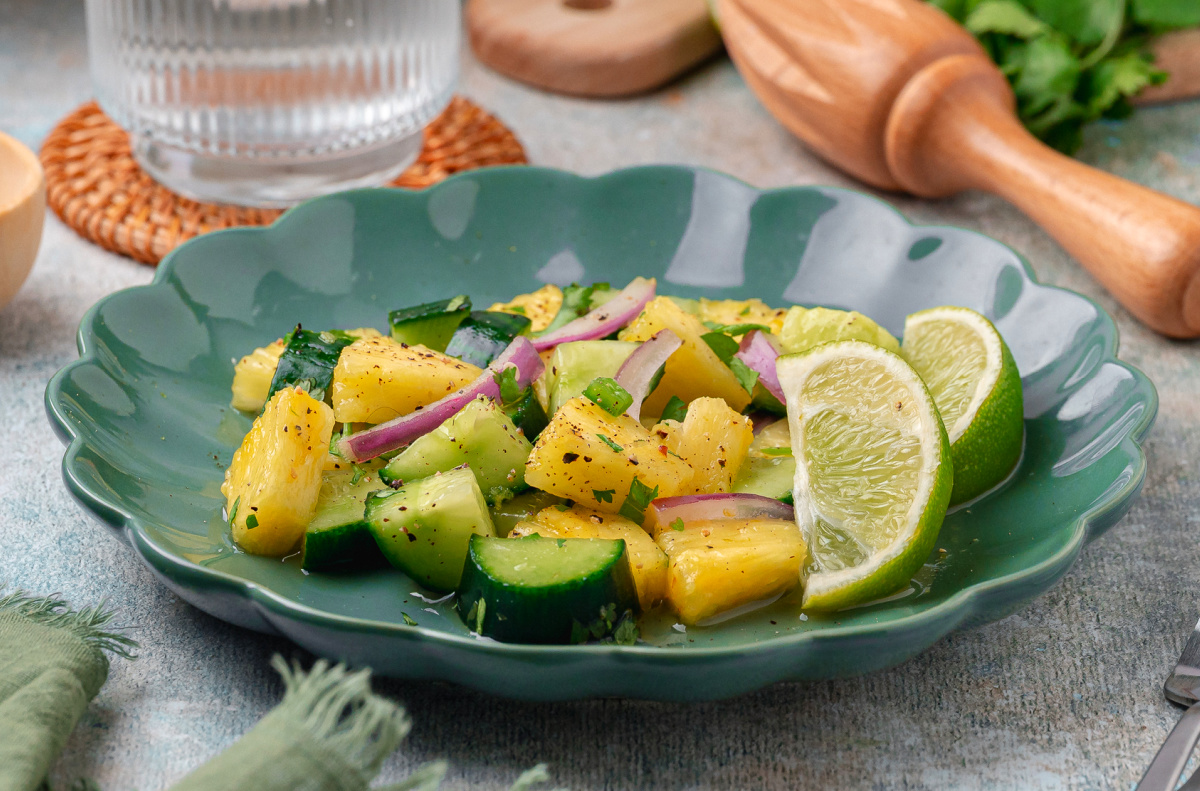 Pineapple and cucumber salad with lime wedges, red onion, and cilantro served on a green ceramic plate.