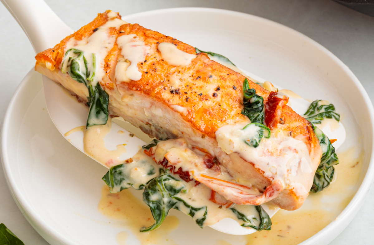 Creamy Tuscan salmon lifted from the pan with a spatula, coated in garlic cream sauce with sun-dried tomatoes and spinach.