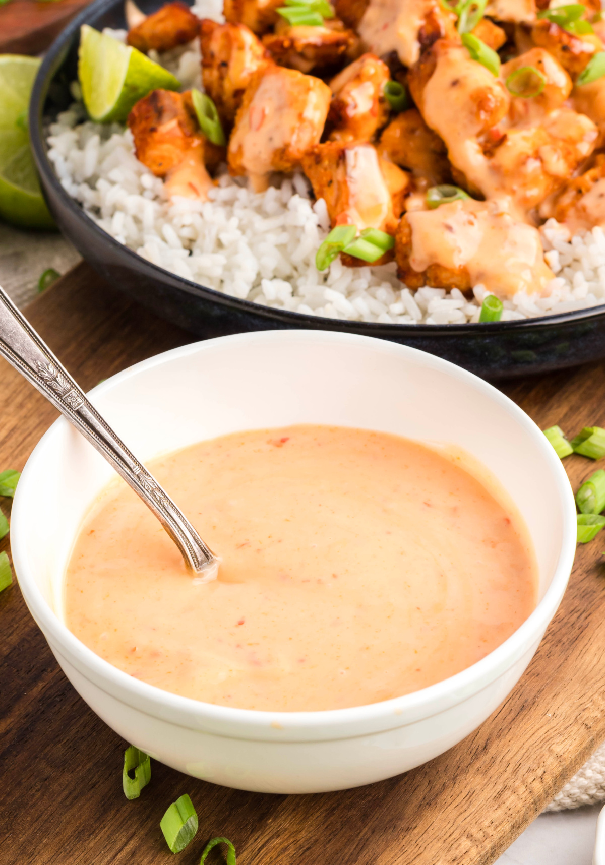 Creamy bang bang sauce in a white bowl with a spoon, served alongside crispy salmon bites over rice in the background.