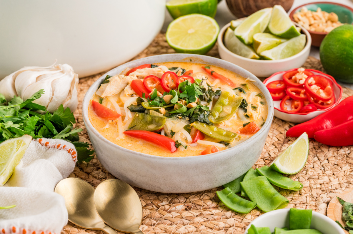 Brothy chicken curry soup with coconut milk, rice noodles, shredded chicken, snow peas, red bell pepper, cilantro, and sliced red chili, served with lime wedges.