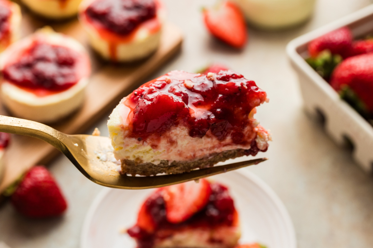 A fork holding a bite of an individual mini cheesecake with graham cracker crust and fresh strawberry topping, showing the creamy interior and layered texture.