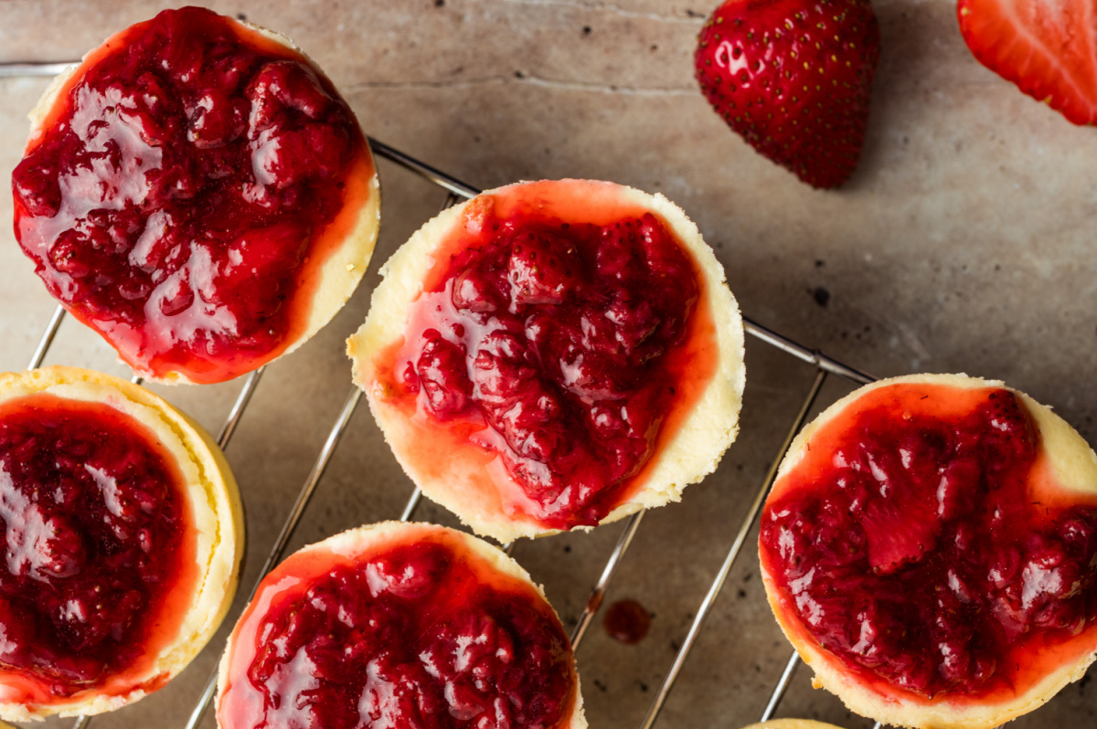 Freshly baked strawberry mini cheesecakes cooling on a wire rack, each topped with a thick layer of cooked strawberry topping.
