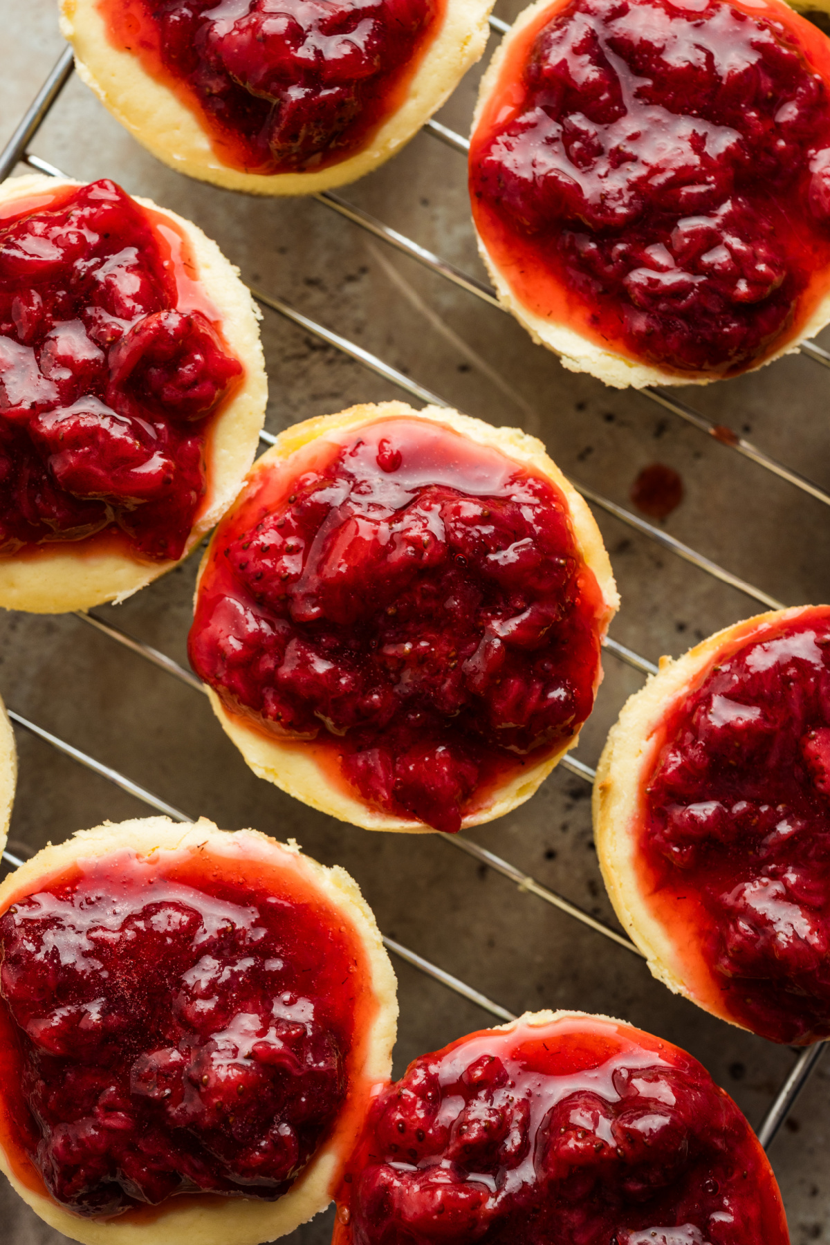 Individual baked mini cheesecakes topped with a glossy strawberry topping, arranged closely together on a wire cooling rack.