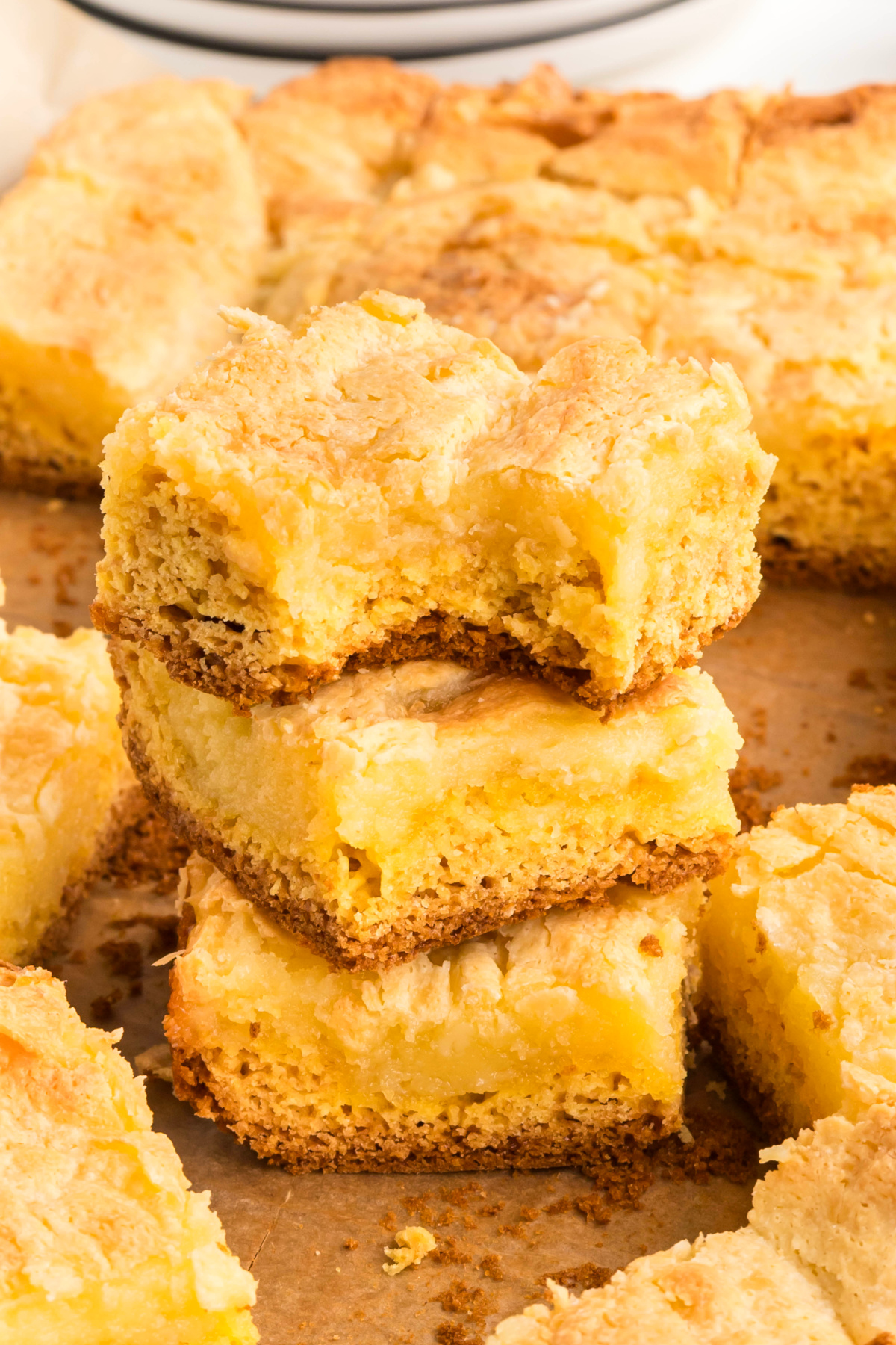 Stacked squares of a cream cheese&ndash;topped cake showing the layered base and soft, custardy center, with crumbs scattered on parchment paper.
