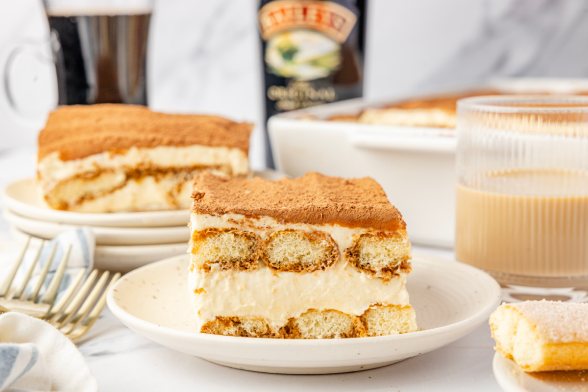 Slice of tiramisu on a plate with visible layers of espresso-soaked ladyfingers and mascarpone cream, with Baileys Irish cream and coffee in the background.