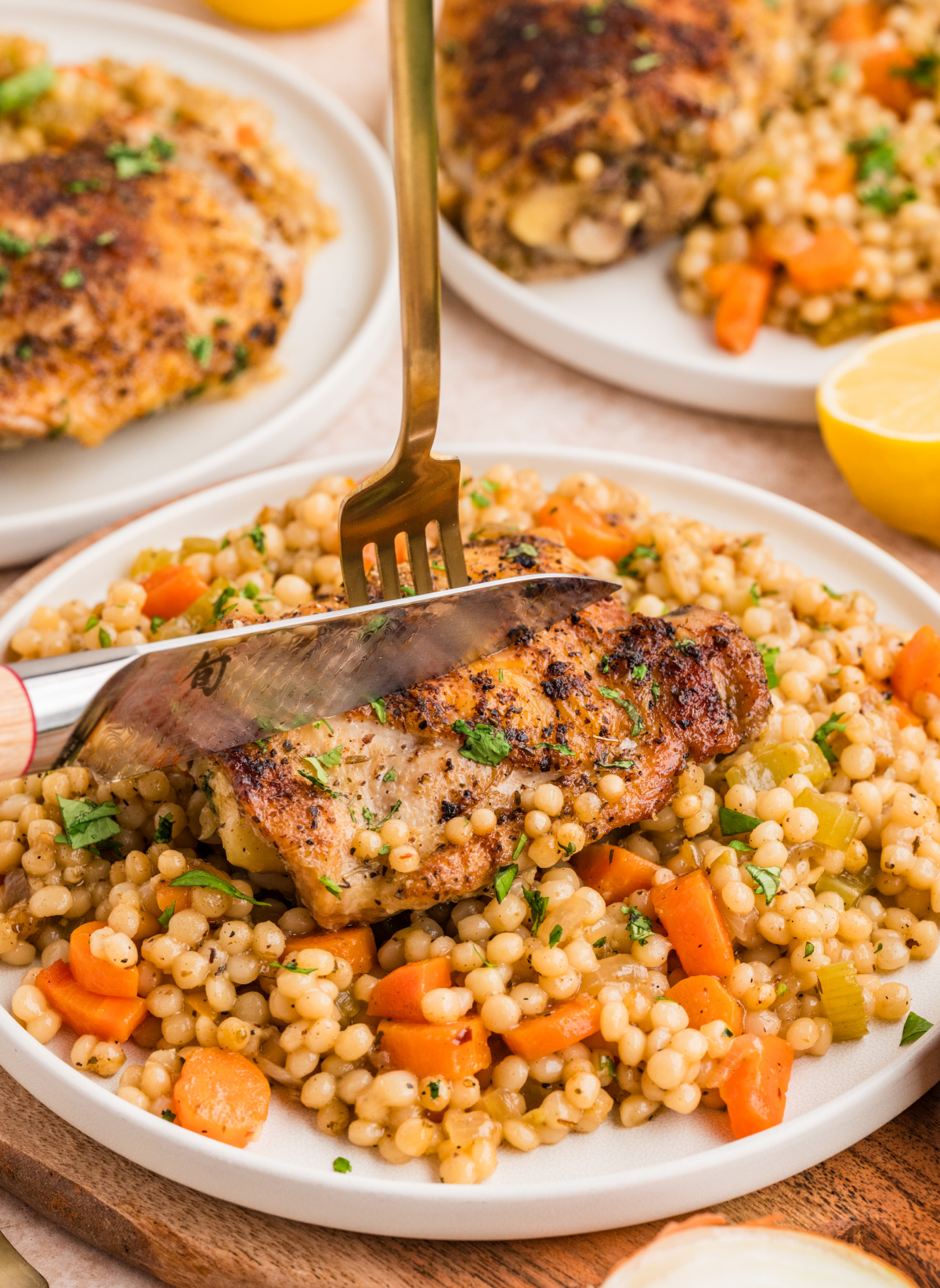 Crispy chicken thighs over pearl couscous served on a plate with carrots and celery, shown being cut with a knife.