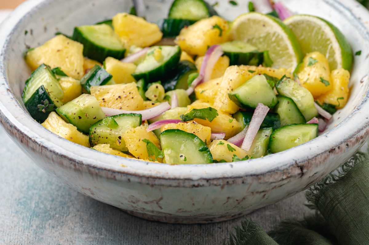 Close-up of pineapple and cucumber salad with red onion, cracked black pepper, cilantro, and lime slices.