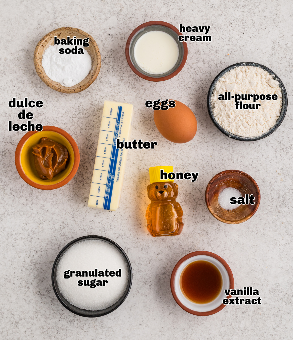 Ingredients for a Russian honey cake laid out on a light surface, including honey, flour, eggs, butter, sugar, dulce de leche, and cream.