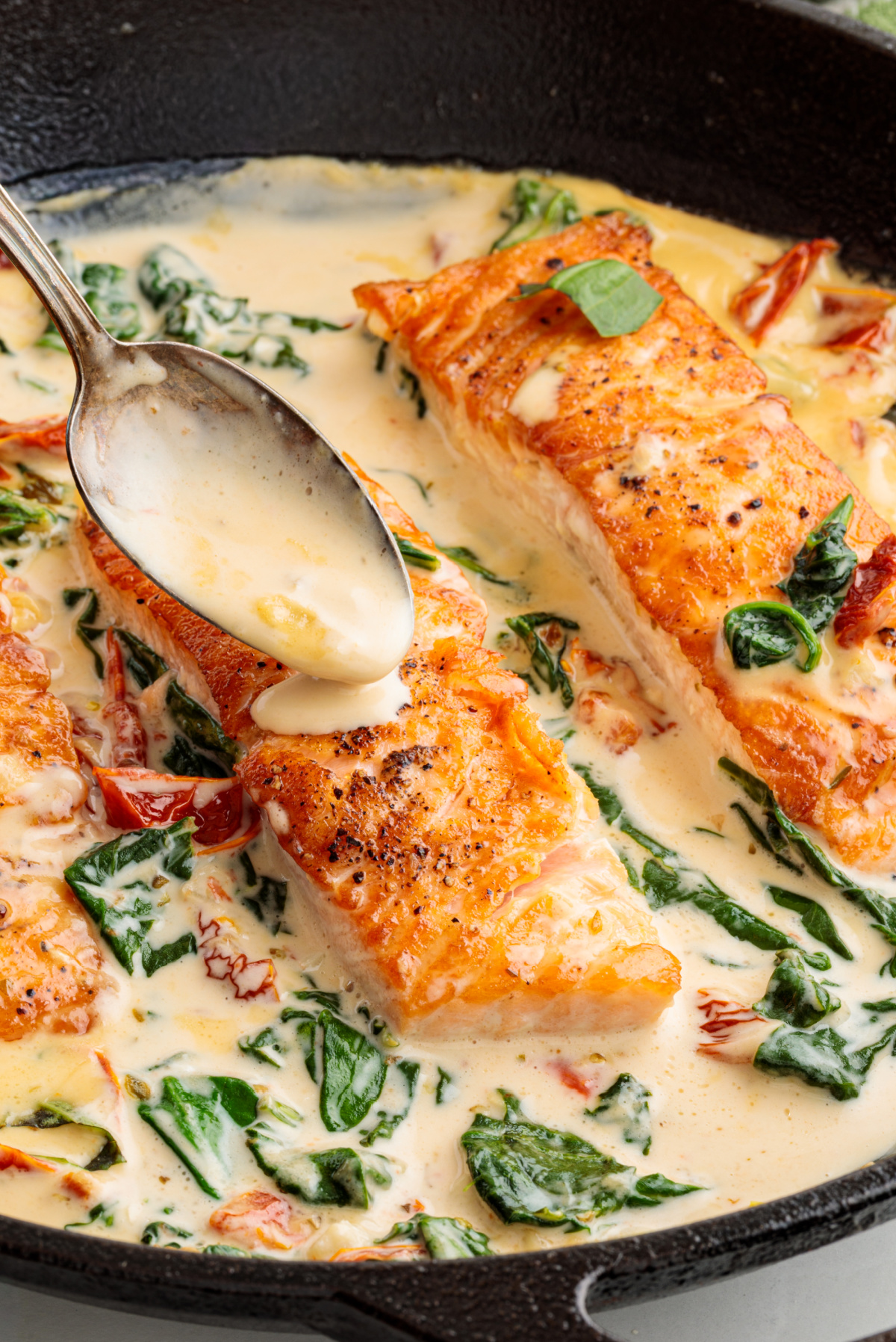 Salmon fillets being spooned with a creamy garlic sauce in a skillet with sun-dried tomatoes and wilted spinach.