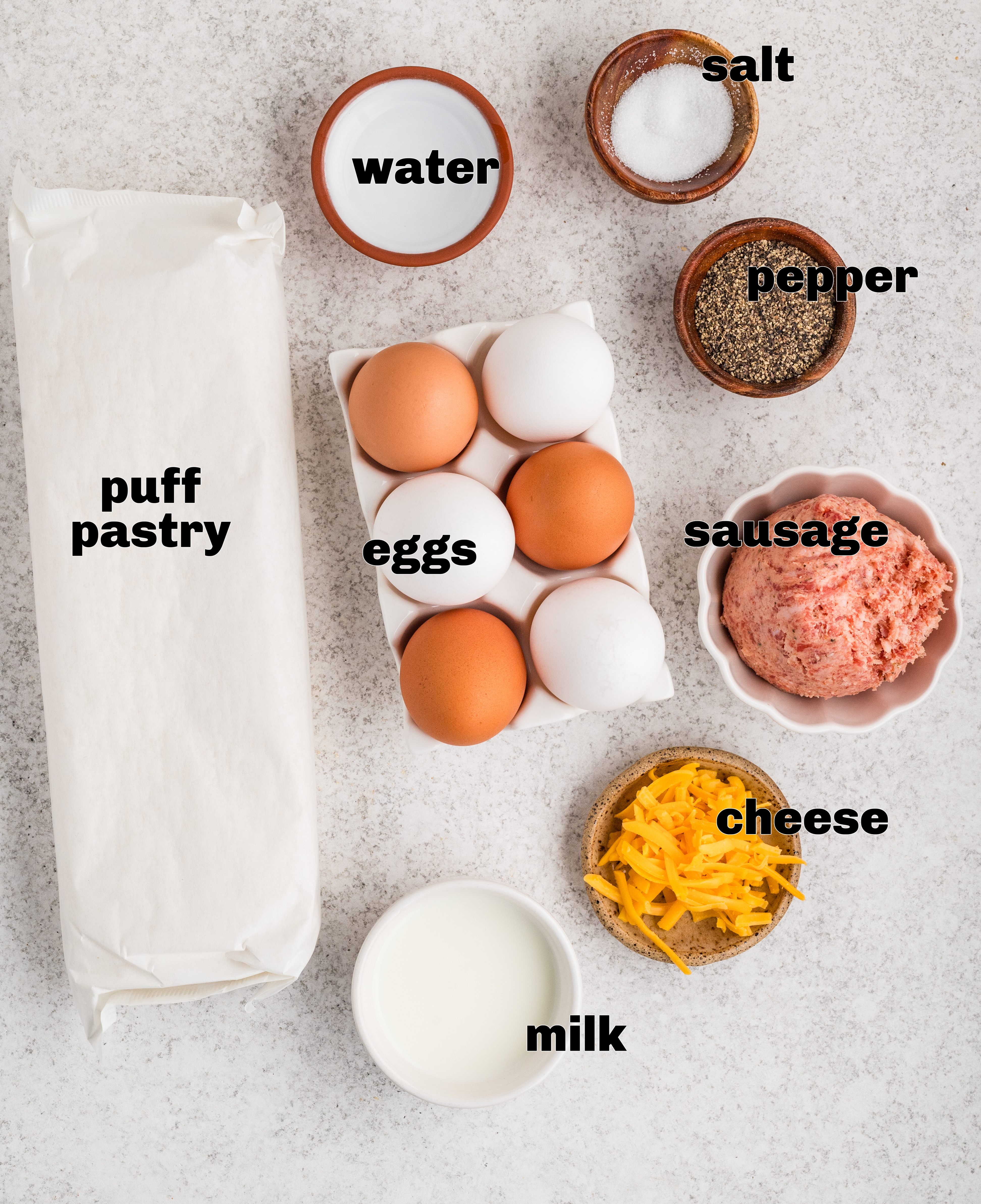 Ingredients for sausage, egg, and cheese breakfast pastries arranged on a light surface, including puff pastry, eggs, sausage, cheddar, milk, salt, and pepper.