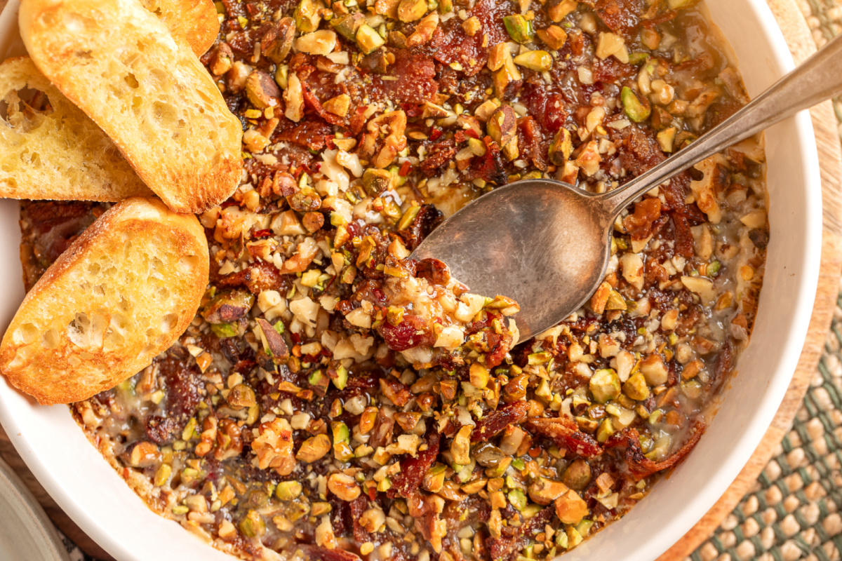 A spoon scooping into a warm baked Boursin dip with bacon, fig preserves, pistachios, walnuts, and toasted crostini on the side.