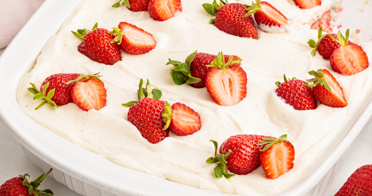 Strawberry Jell-O poke cake in a white baking dish topped with whipped vanilla cream cheese frosting and fresh strawberry halves.