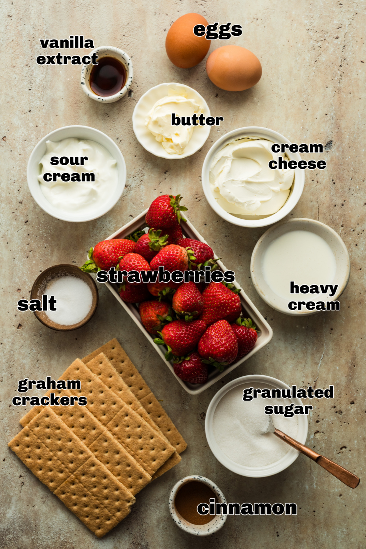 The ingredients for strawberry mini cheesecakes arranged on a countertop, including graham crackers, cream cheese, fresh strawberries, eggs, sugar, butter, sour cream, heavy cream, vanilla, cinnamon, and salt.