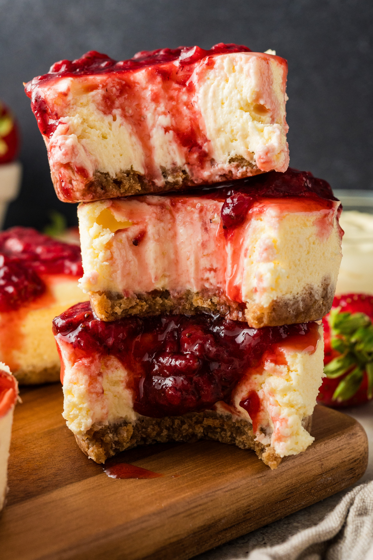 Three strawberry mini cheesecakes stacked on a wooden board, showing the creamy cheesecake interior, graham cracker crust, and thick strawberry topping dripping down the sides.