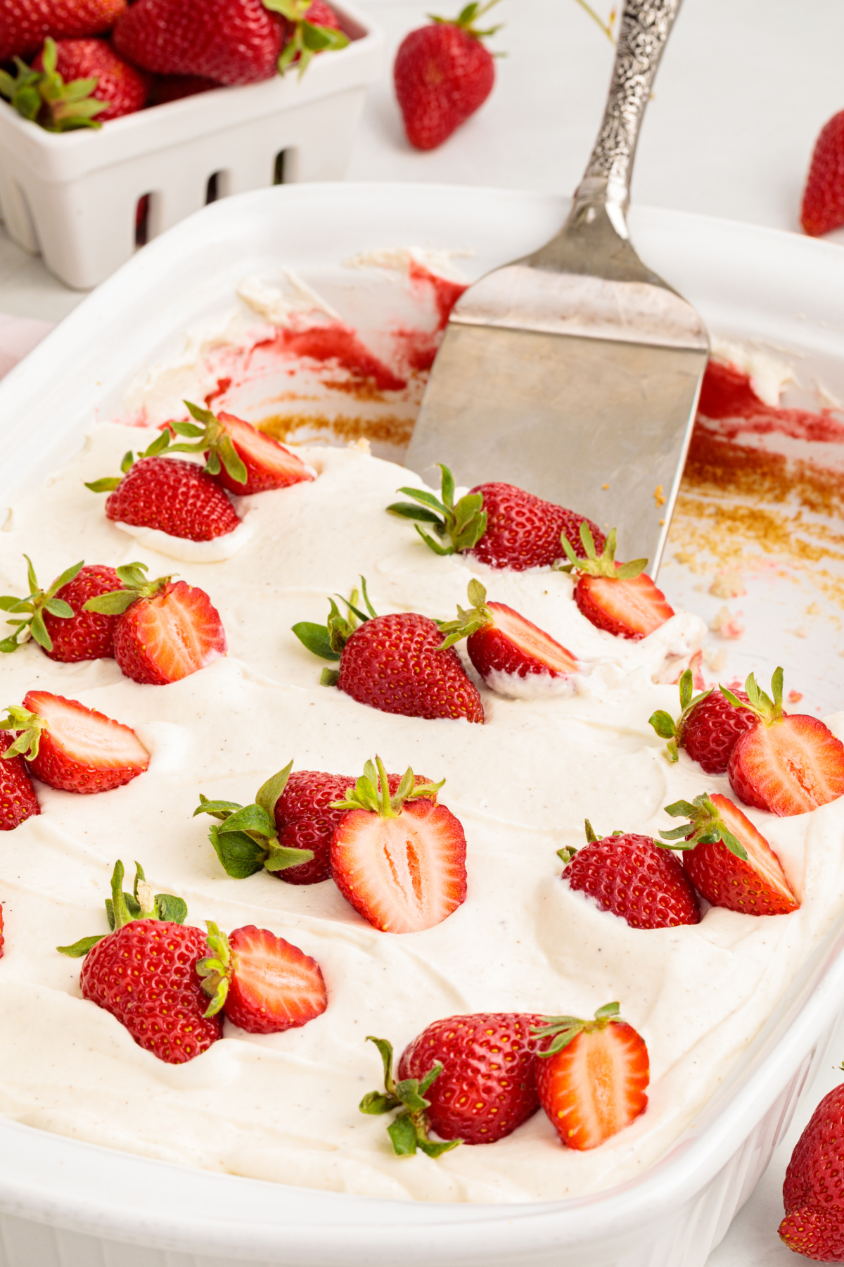 Strawberry poke cake in a baking dish with whipped vanilla cream cheese frosting spread smoothly on top and finished with fresh strawberry halves.