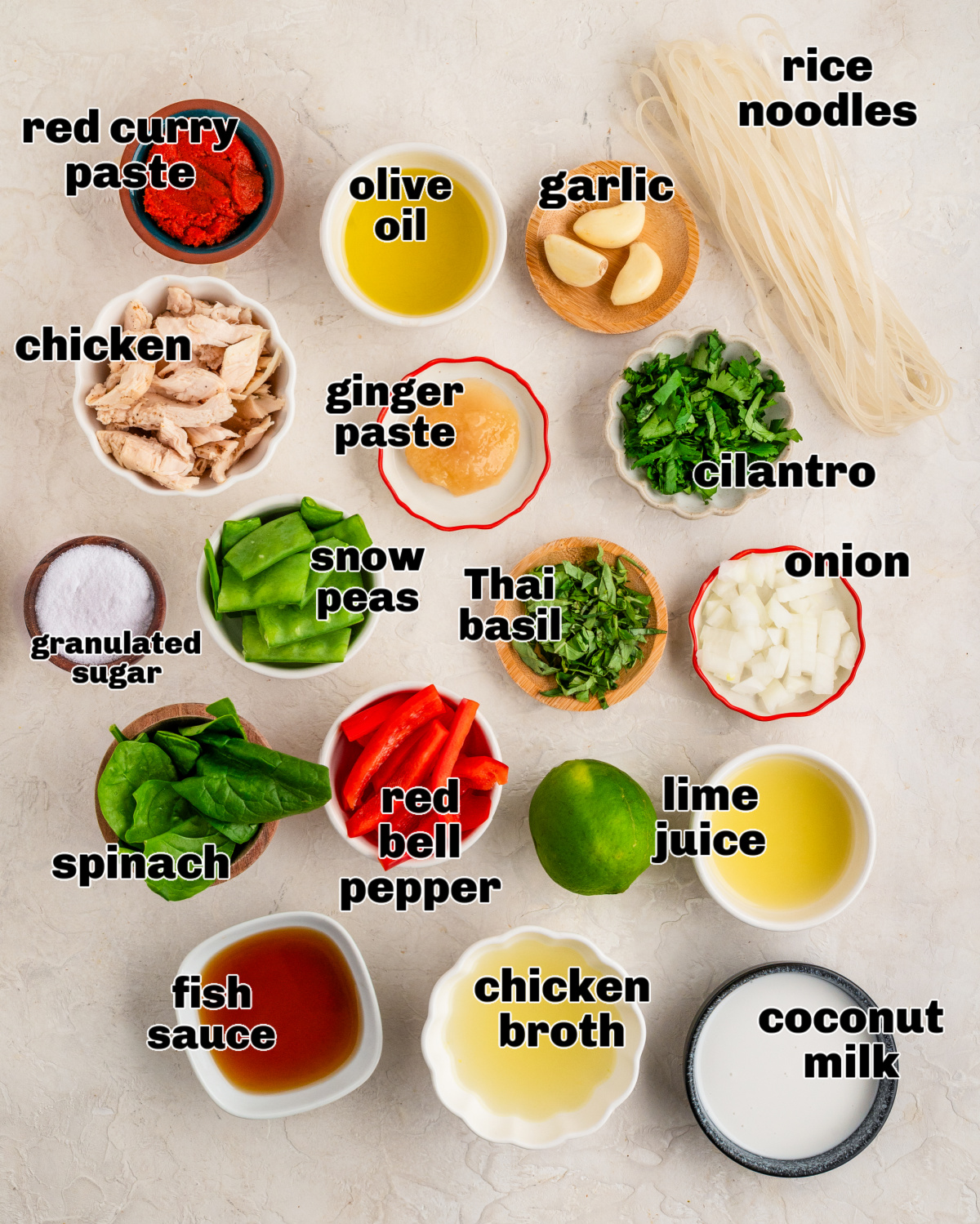 Ingredients for coconut curry chicken soup arranged on a light surface, including red curry paste, shredded chicken, rice noodles, coconut milk, chicken broth, snow peas, spinach, red bell pepper, onion, garlic, ginger paste, lime juice, fish sauce, sugar, cilantro, and Thai basil.