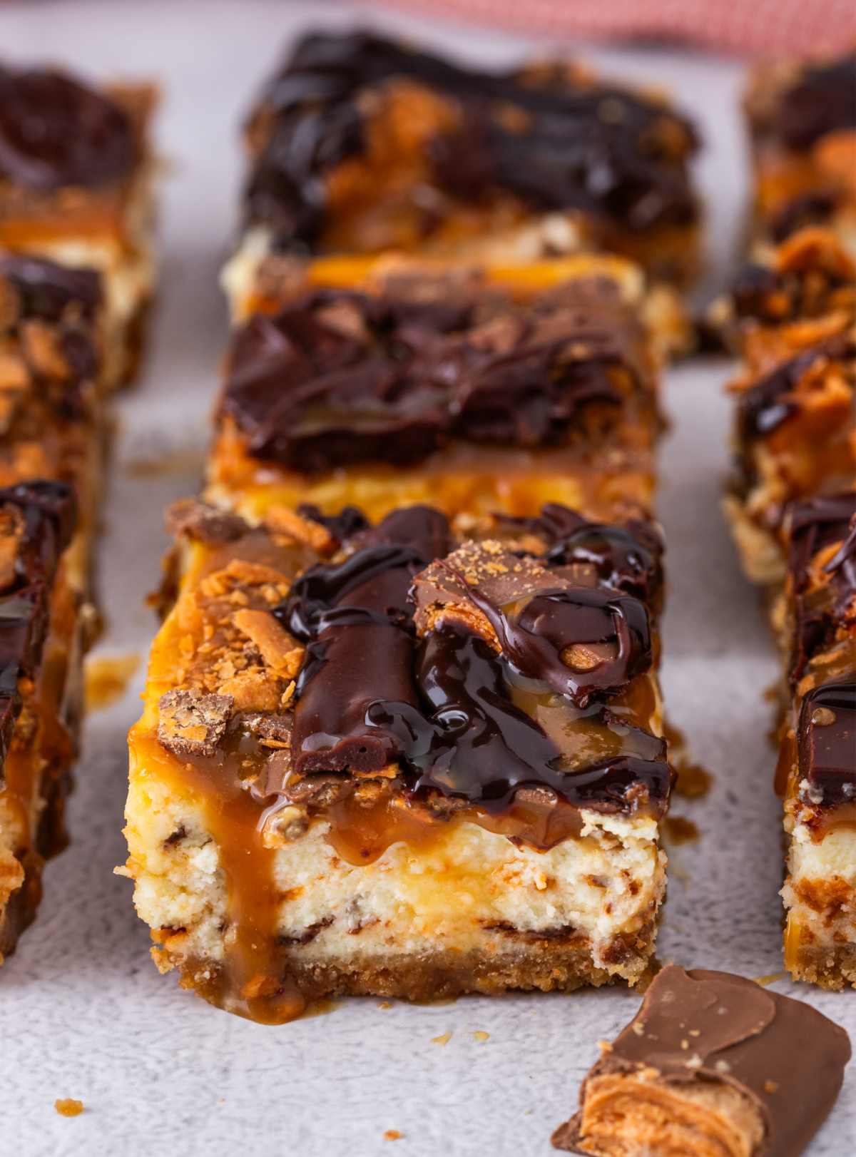 Caramel cheesecake bars topped with chopped Butterfinger candy and chocolate drizzle