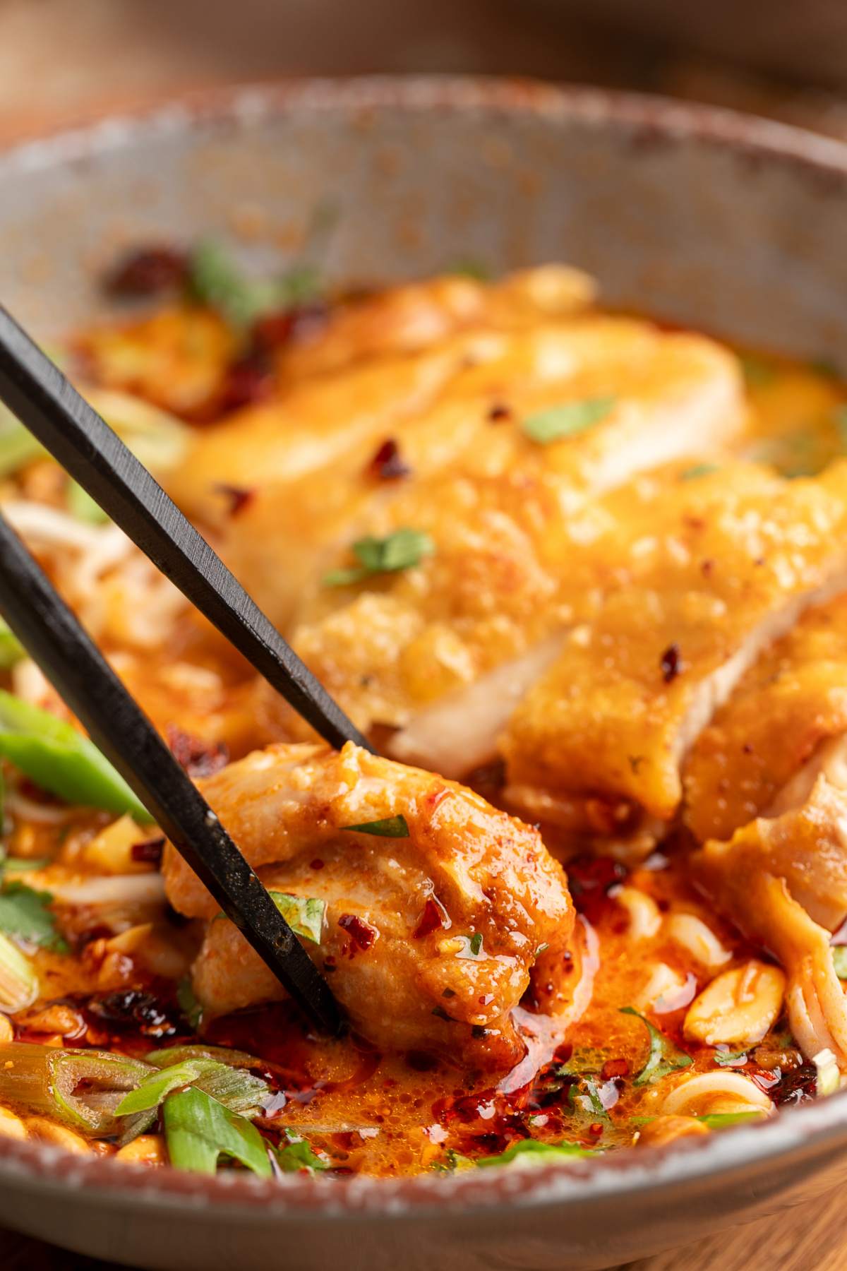 Chopsticks lifting a piece of crispy chicken from Thai coconut curry ramen with chili oil, scallions, and peanuts