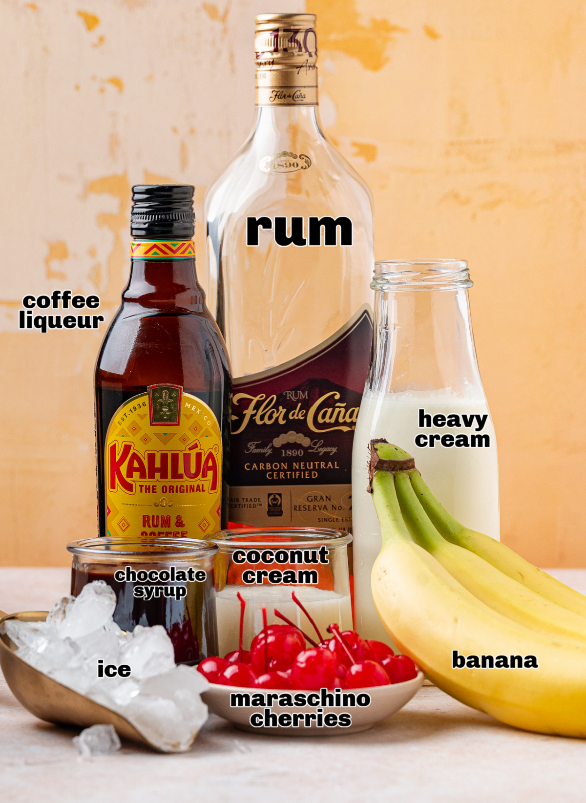 Ingredients for banana rum cocktail including dark rum, coffee liqueur, heavy cream, cream of coconut, chocolate syrup, ice, and maraschino cherries