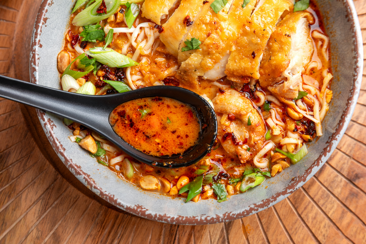 Spoonful of coconut broth from panang curry ramen with crispy chicken, chili oil, scallions, and peanuts