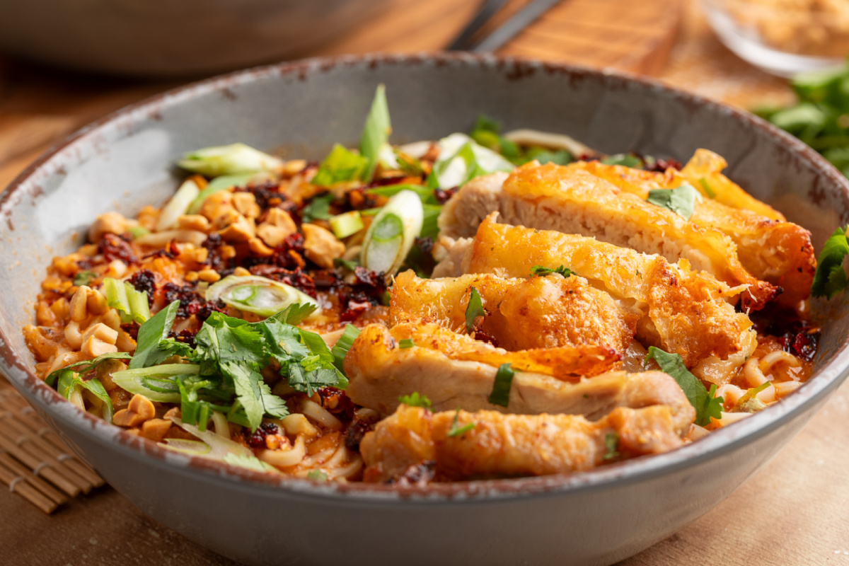 Close-up of panang curry ramen with crispy chicken, coconut broth, chili crunch, scallions, and chopped peanuts