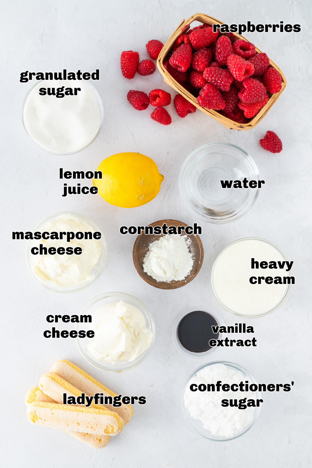 Ingredients for raspberry tiramisu arranged on a white surface, including raspberries, mascarpone, cream cheese, and ladyfingers