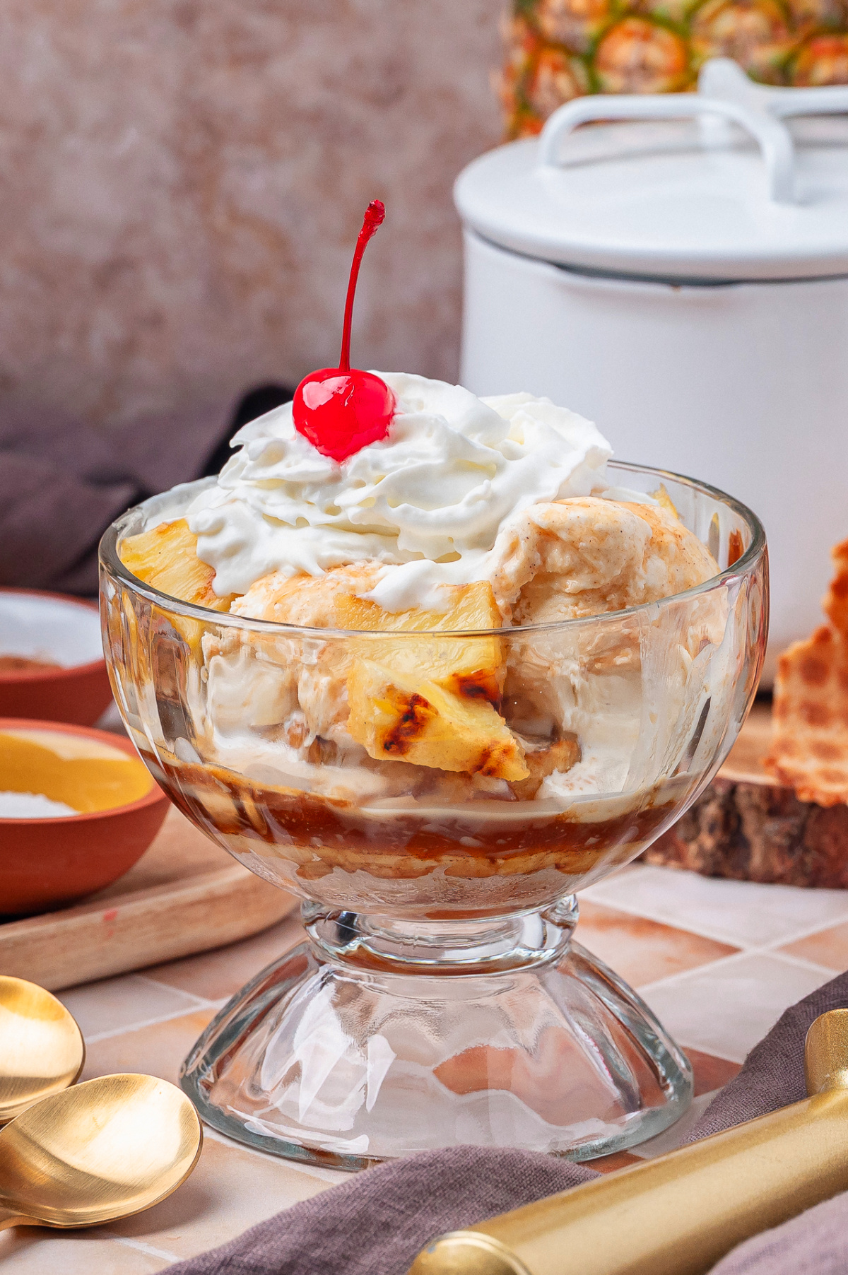 Rum glazed grilled pineapple with vanilla ice cream, whipped cream, and cherry in a glass dessert bowl