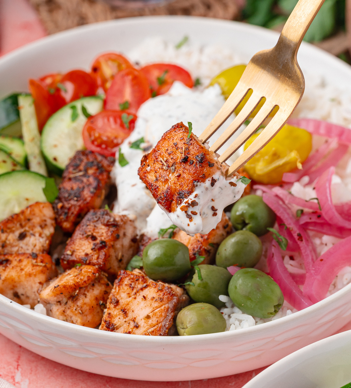 Seared salmon cube with yogurt dill sauce over white rice, olives, pickled red onions, and fresh vegetables in a Mediterranean salmon bowl