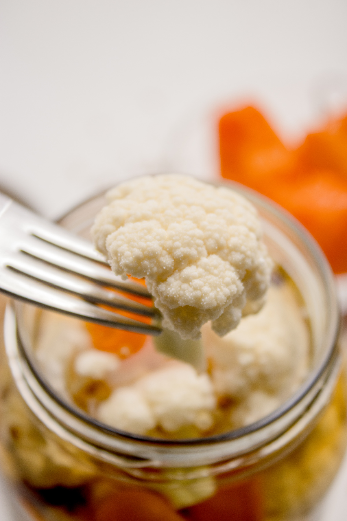 Fork lifting a crisp cauliflower floret from spicy vinegar brine in glass jar