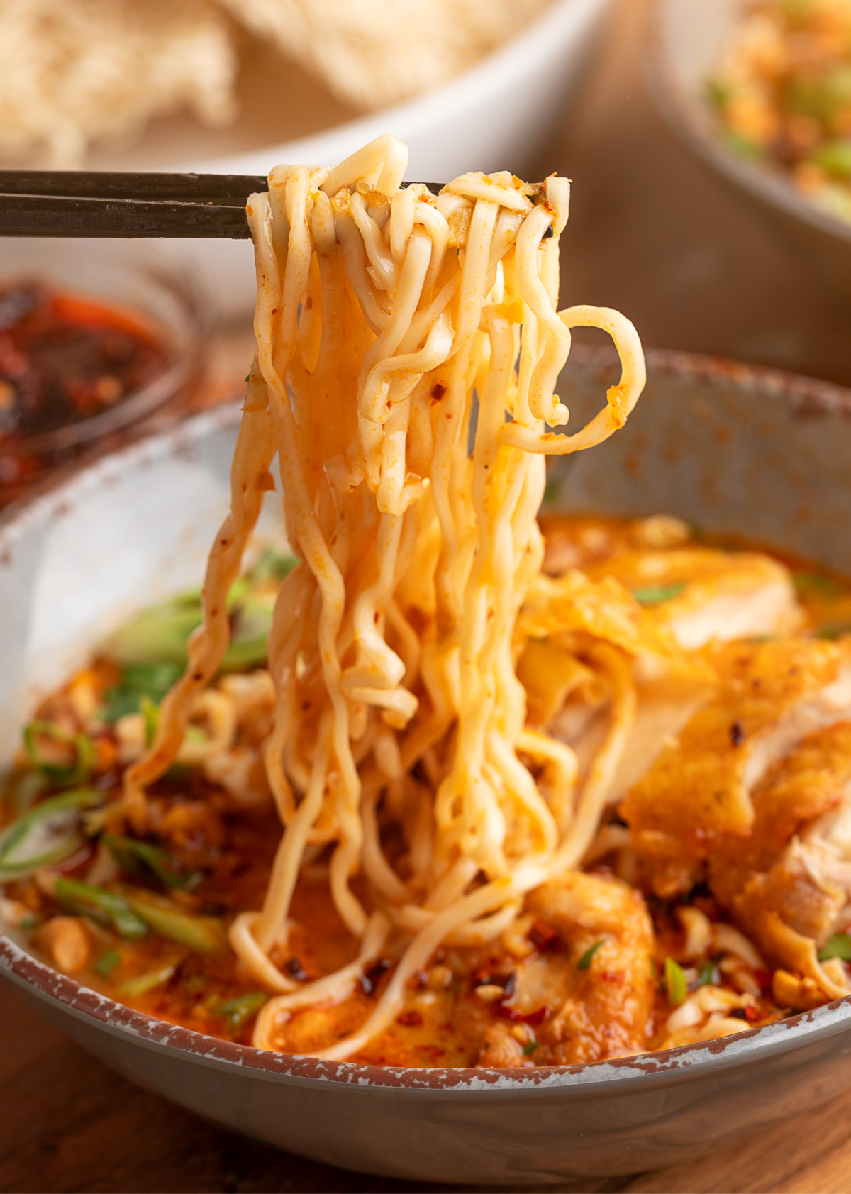Chopsticks lifting ramen noodles from Thai coconut curry ramen with crispy chicken, chili oil, and scallions