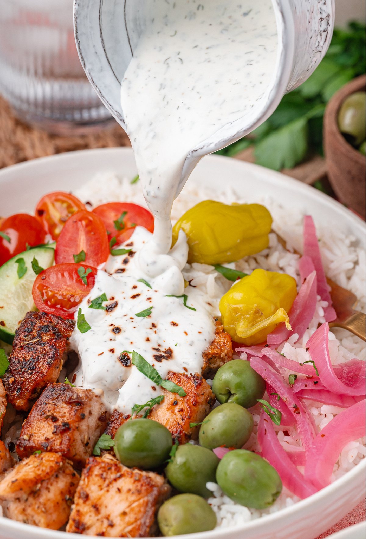 Yogurt dill sauce being poured over seared salmon, white rice, olives, and pickled red onions in a Mediterranean bowl