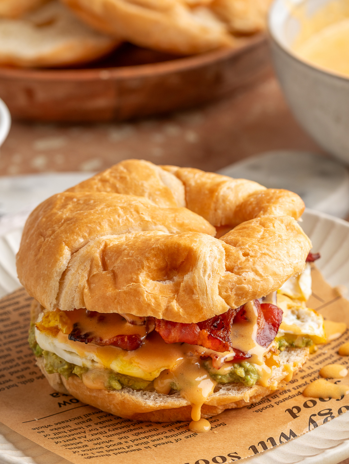 Bacon egg avocado breakfast sandwich stacked on a flaky croissant with crispy bacon, fried egg and lime chipotle hollandaise dripping down the side