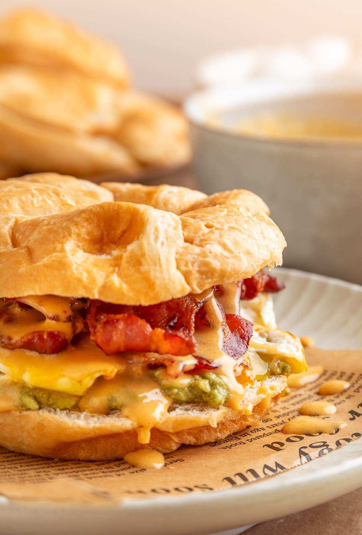 Close side view of a bacon egg avocado breakfast sandwich on a croissant with melted yolk, crispy bacon and creamy chipotle hollandaise