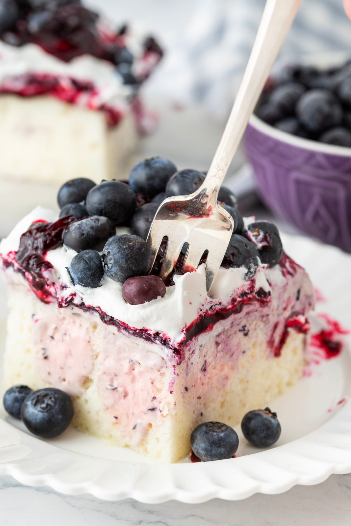 Fork cutting into blueberry cheesecake poke cake with fresh blueberries and whipped topping