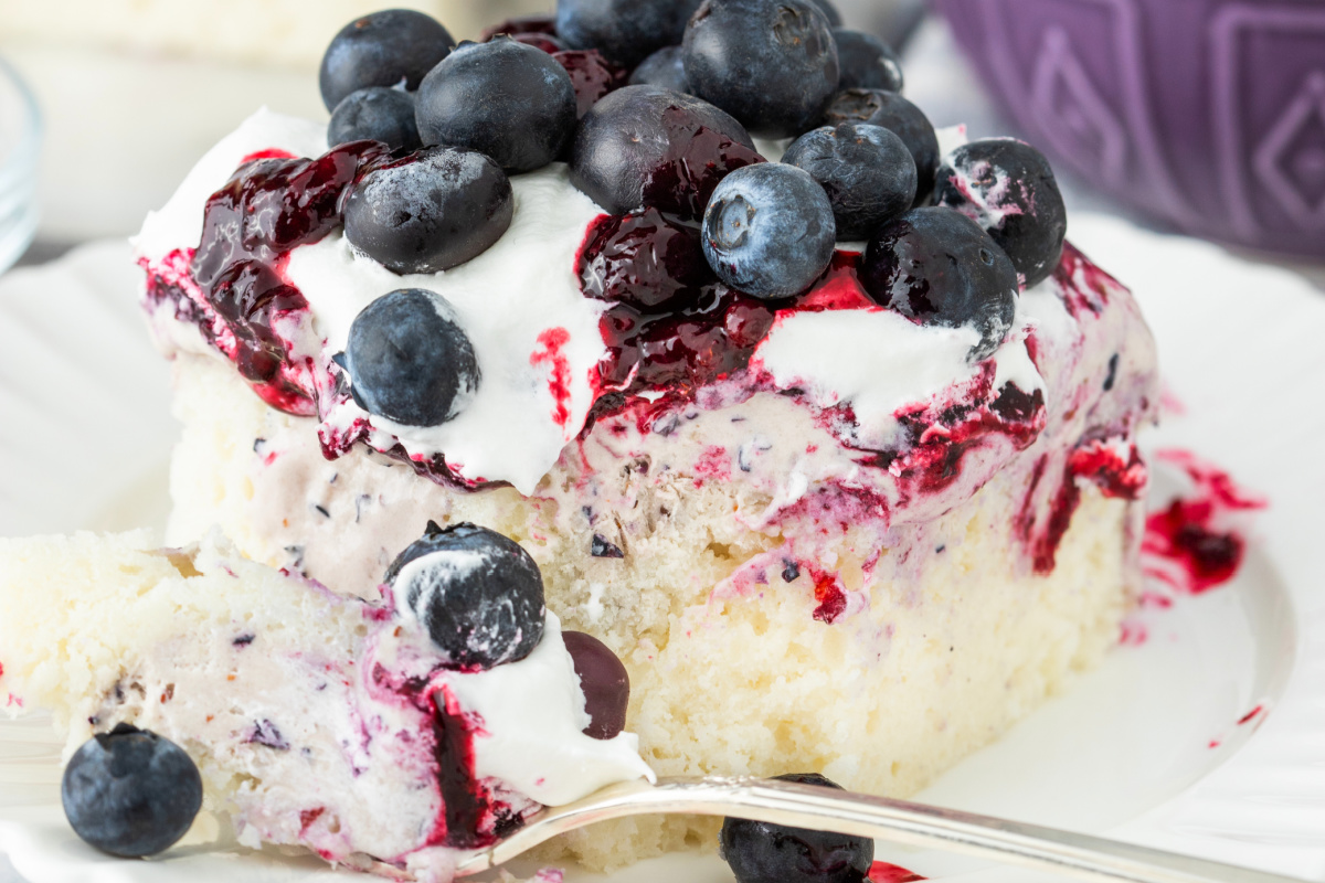 Slice of blueberry poke cake topped with whipped topping, blueberry compote, and fresh blueberries