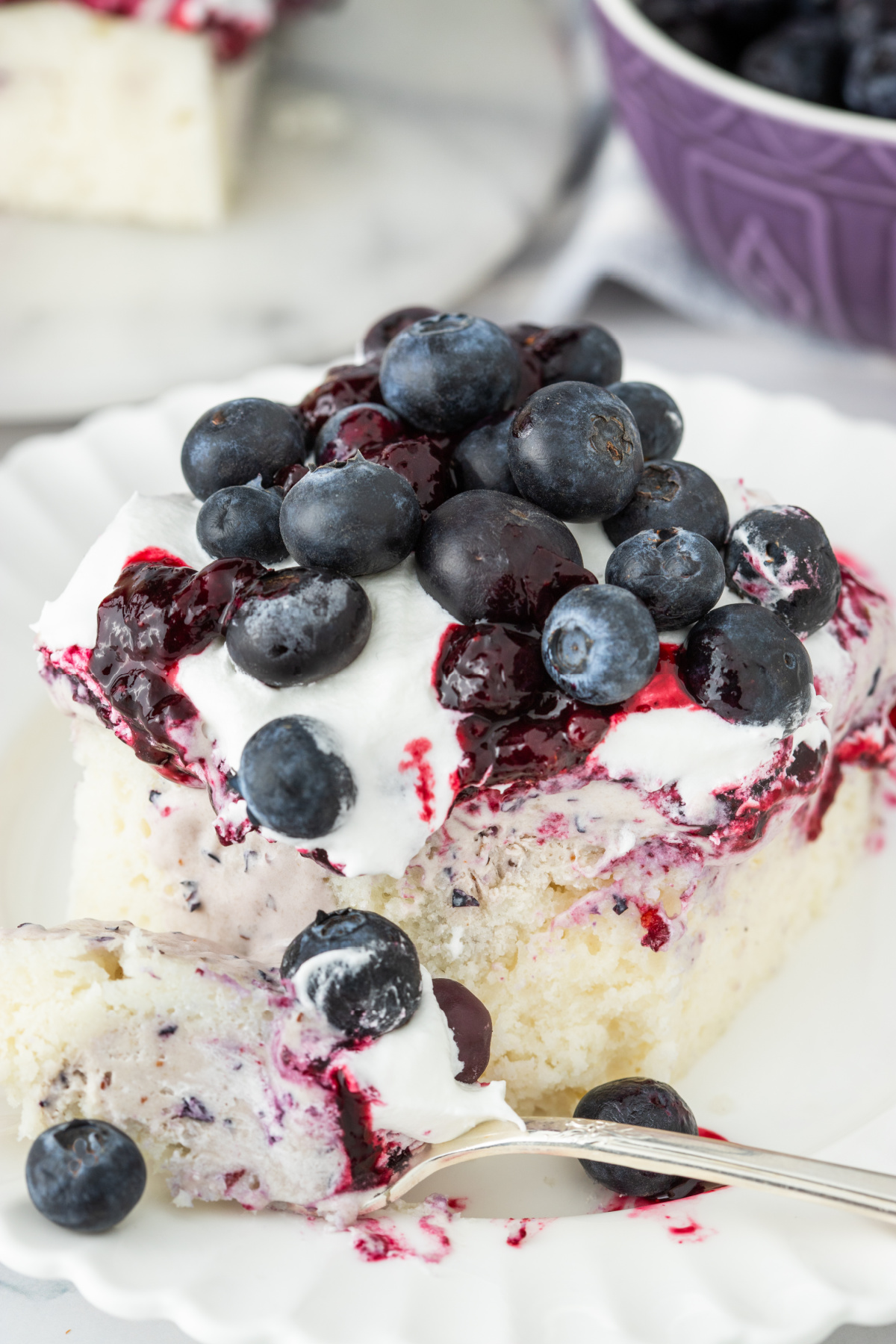 Slice of blueberry poke cake with creamy filling, blueberry topping, and fresh berries