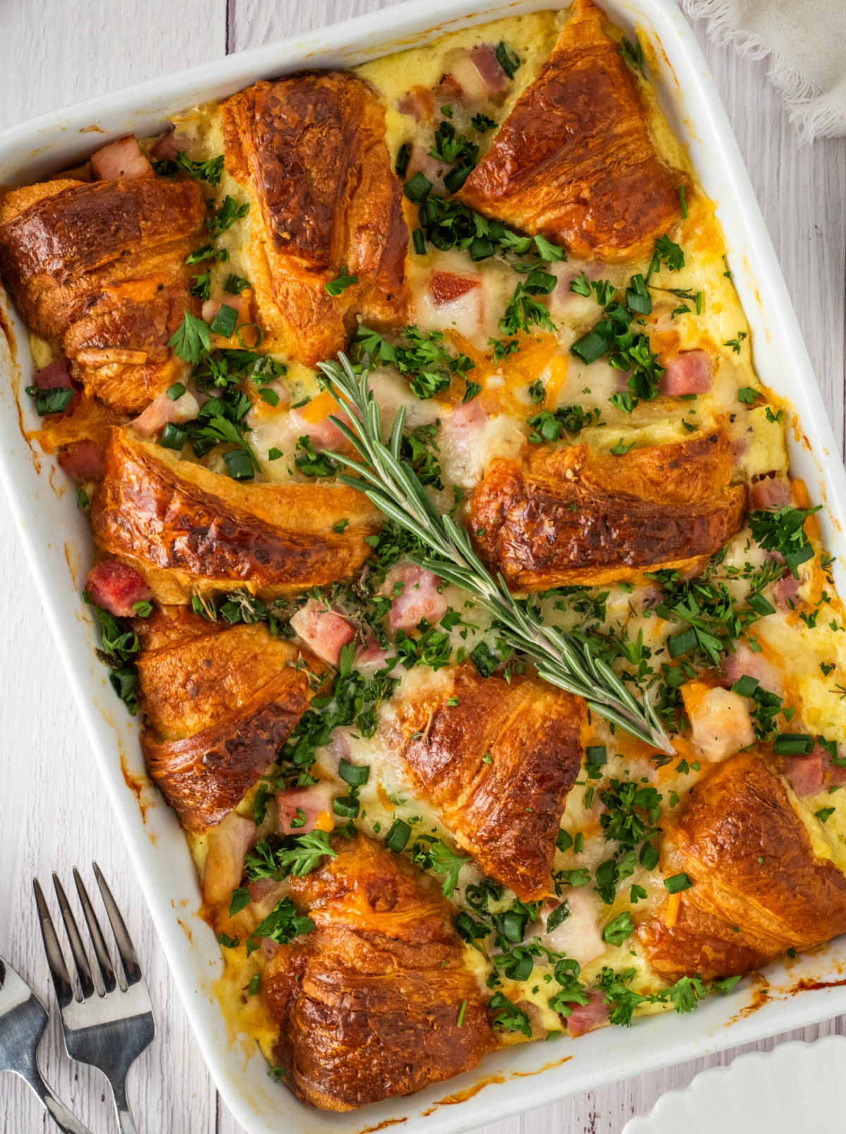 golden croissant breakfast bake with ham, melted Swiss and cheddar cheese, and fresh herbs baked in a white casserole dish
