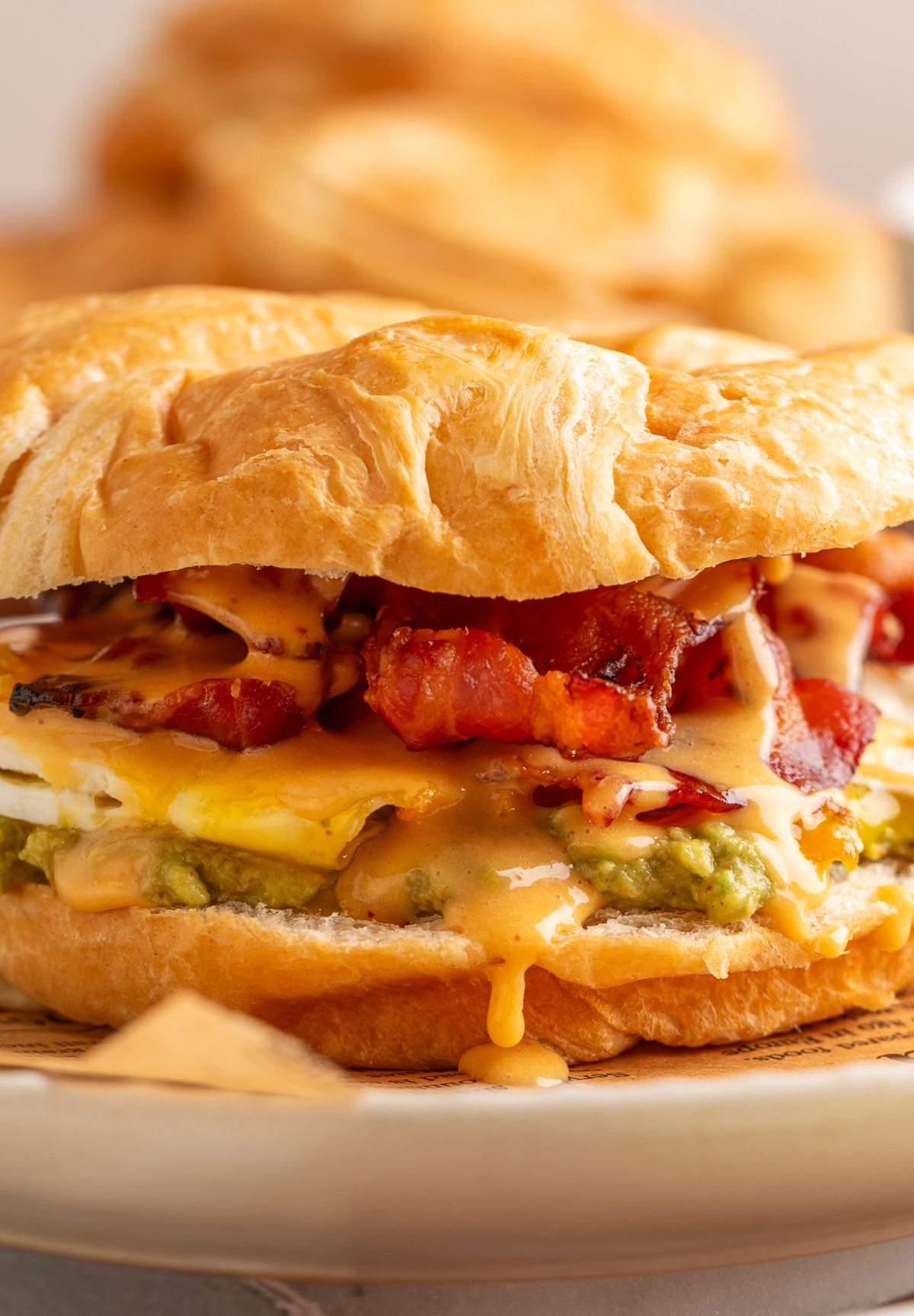 Close up of croissant breakfast sandwich layers with mashed avocado, fried egg, crispy bacon and spicy chipotle hollandaise dripping