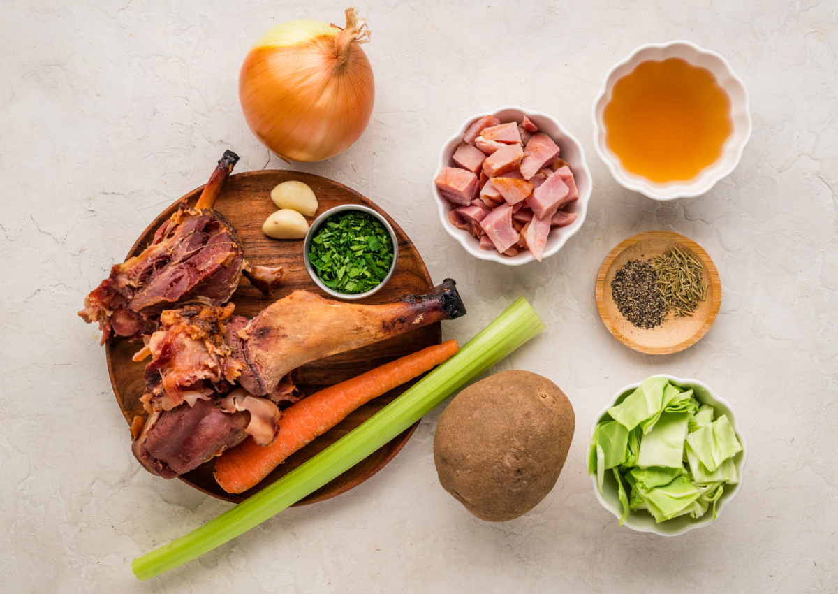 Ingredients for ham and cabbage soup including ham hock, cabbage, potatoes, carrots, celery, onion, garlic, and broth