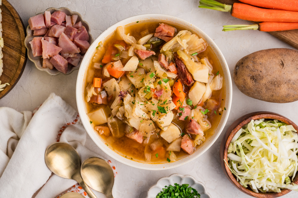 Ham and cabbage soup with potatoes, carrots, cabbage, and ham in a brothy bowl with ingredients around it