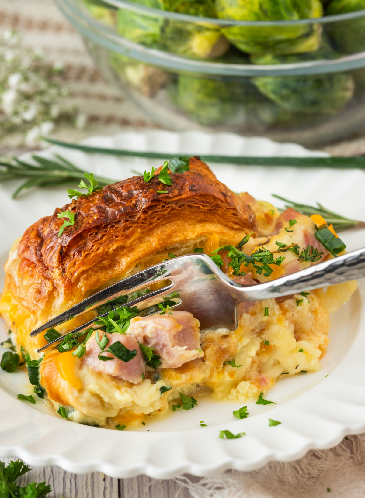 slice of croissant breakfast bake with ham, melted cheese, and egg custard on a white plate with a fork