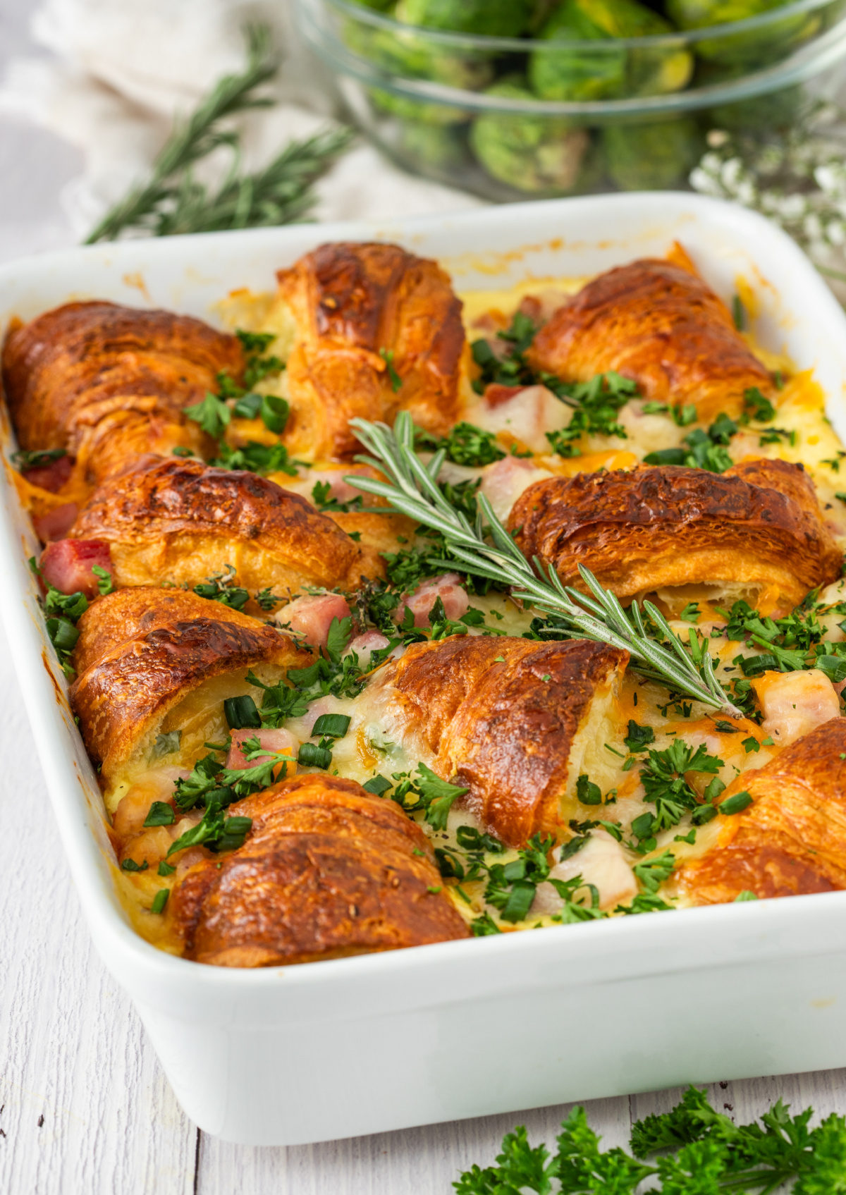 ham and cheese croissant breakfast bake with golden croissants, diced ham, melted cheese, and fresh herbs baked in a white casserole dish