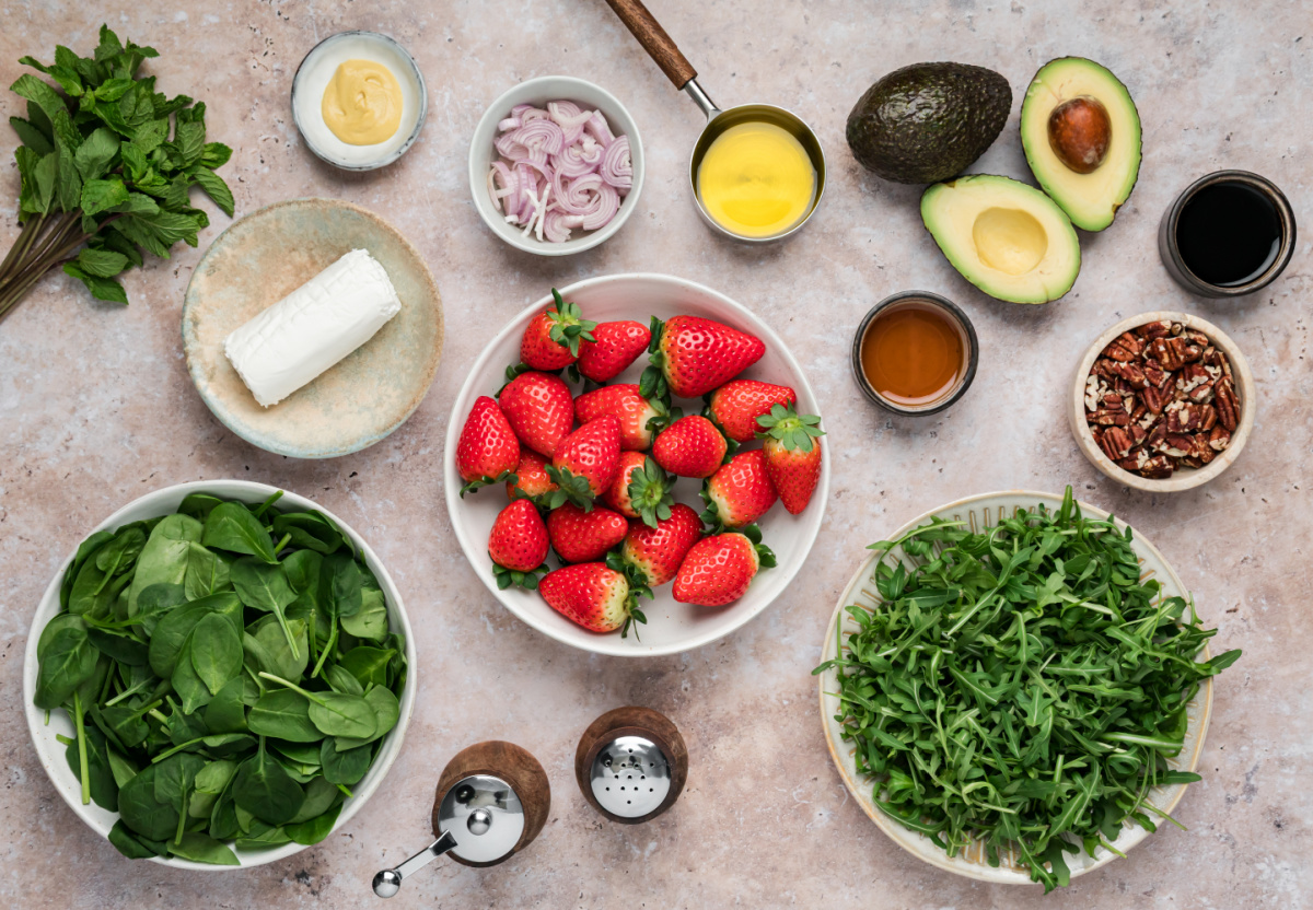 Ingredients for strawberry salad with goat cheese including fresh strawberries, spinach, arugula, avocado, pecans, and balsamic vinaigrette