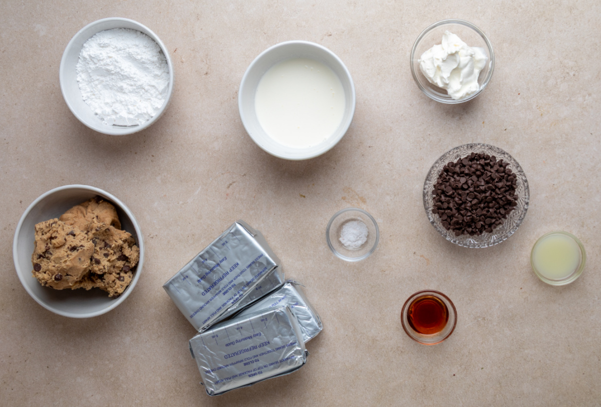 ingredients for cookie dough crust cheesecake including cream cheese, cookie dough, powdered sugar, cream, and chocolate chips