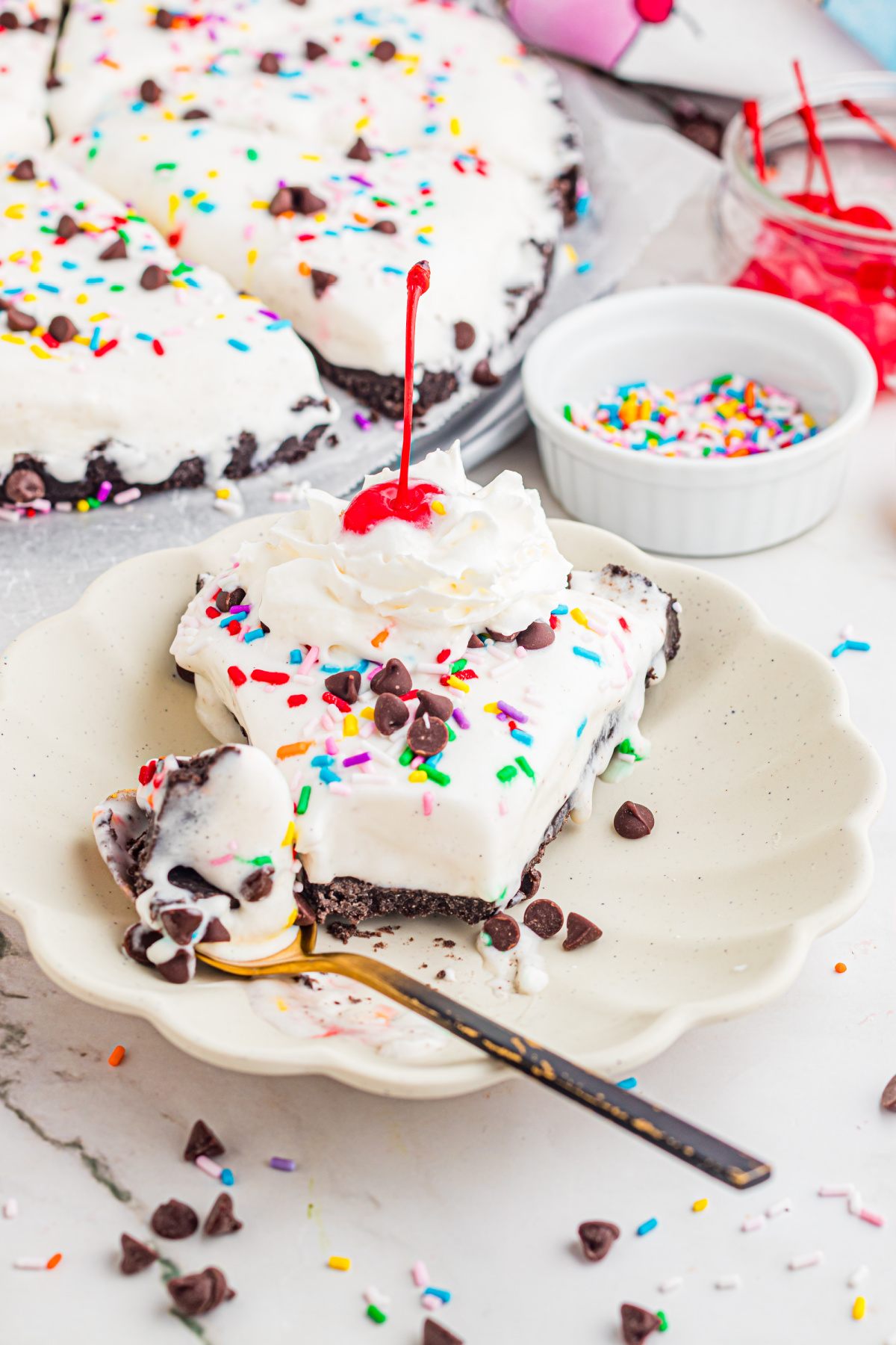 slice of ice cream dessert pizza with Oreo crust topped with whipped cream, cherry, sprinkles, and chocolate chips