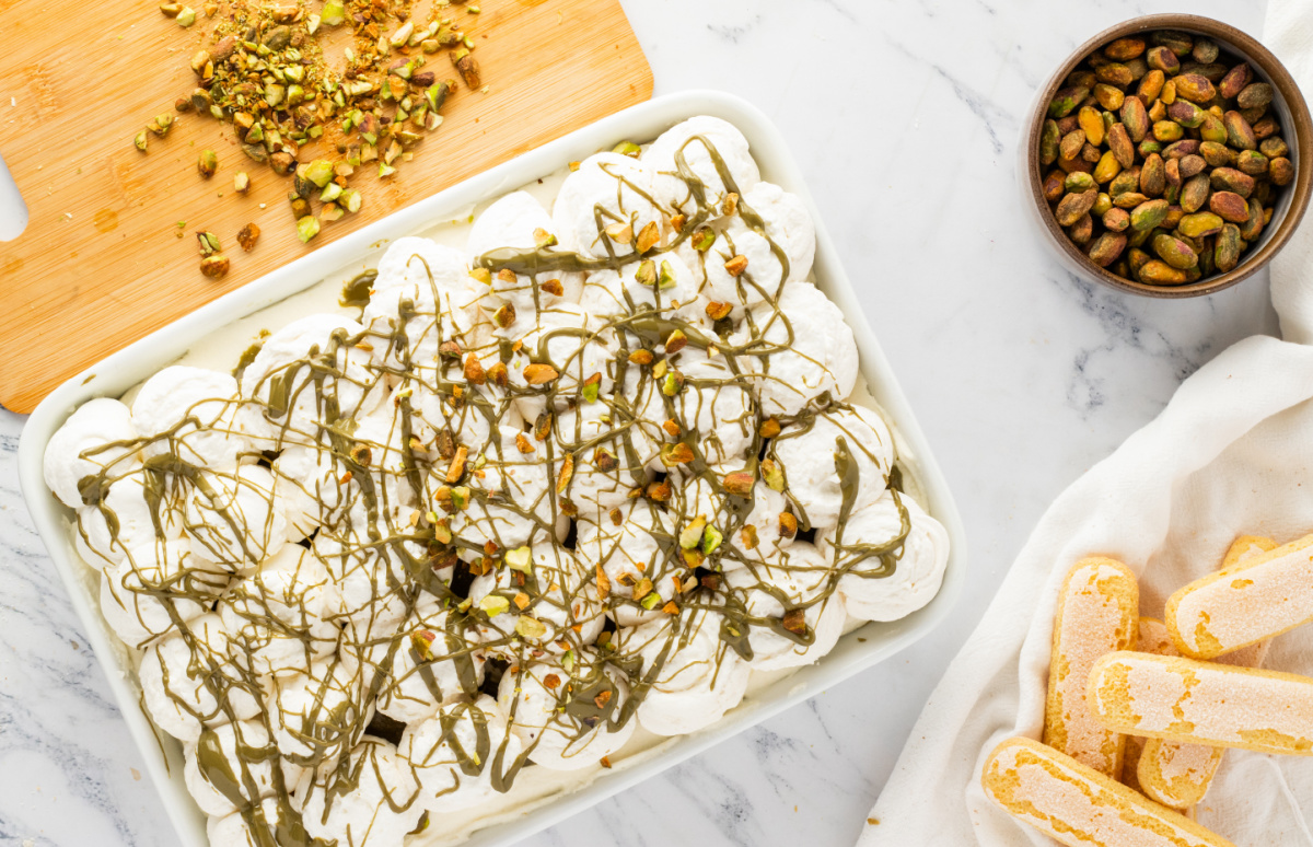 pistachio tiramisu in a 9x13 pan topped with whipped cream pistachio drizzle and chopped pistachios with ladyfingers and pistachios on the side