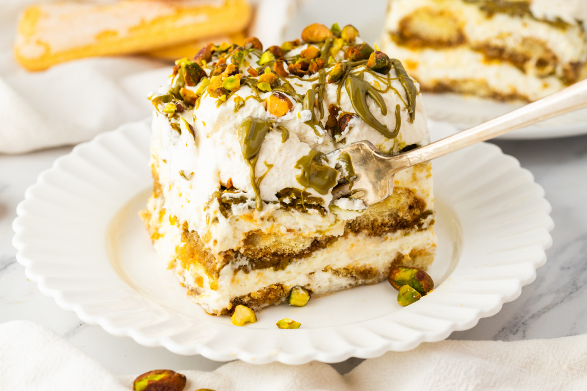 pistachio tiramisu with fork cutting into layers of mascarpone cream and espresso soaked ladyfingers topped with pistachio drizzle and chopped pistachios